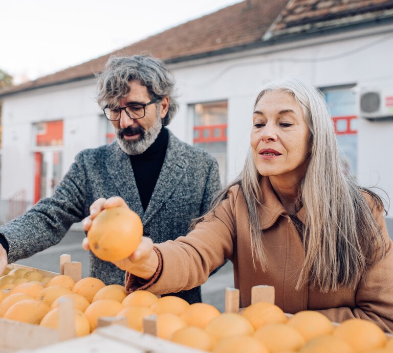  Ein älteres Paar mit grauen Haaren steht an einem Obststand und wählt gemeinsam frische Orangen aus. Der Mann, der eine Brille und einen grauen Wollmantel trägt, hält eine Orange hoch, während die Frau mit langen, glatten Haaren und einem braunen Mantel aufmerksam zusieht. Sie scheinen die Qualität der Früchte zu begutachten, was auf ein Bewusstsein für gesunde Ernährung hinweist. Im Hintergrund ist ein Teil eines Gebäudes mit einem roten Fensterrahmen zu sehen, was auf ein öffentliches Umfeld wie einen Markt hinweisen könnte. | © Getty Images/RgStudio