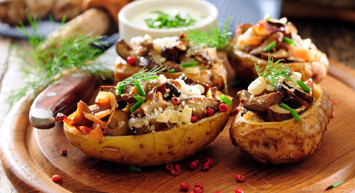 Gebackene Kartoffelhälften mit Pilzen, Karotten und Parmesan liegen mit Preiselbeeren und Schnittlauch angerichtet auf einem runden Holzbrett. Im Hintergrund liegt ein Messer, mehrere Pilze und eine Schale mit einem Kräuterdip.