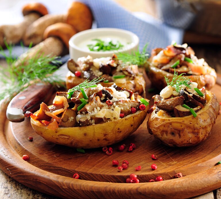 Gebackene Kartoffelhälften mit Pilzen, Karotten und Parmesan liegen mit Preiselbeeren und Schnittlauch angerichtet auf einem runden Holzbrett. Im Hintergrund liegt ein Messer, mehrere Pilze und eine Schale mit einem Kräuterdip. | © Shutterstock / zebratomato