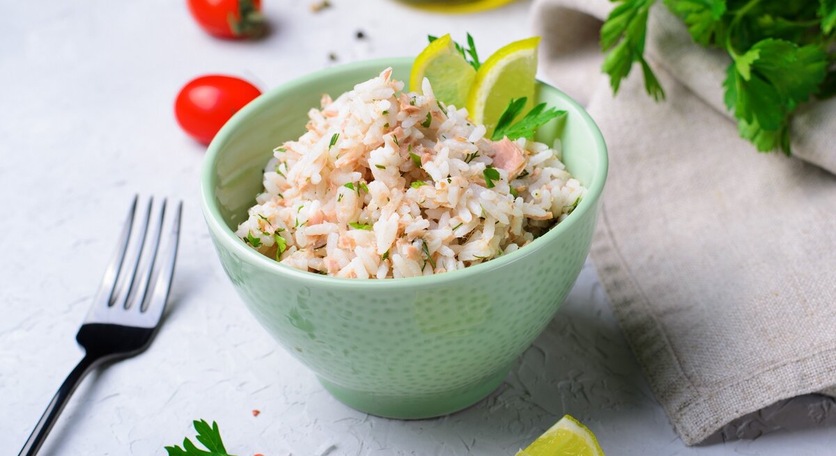 Eine kleine grüne Schale mit dem Thunfisch-Reis auf einem grauen Tisch mit mausgrauer Decke. Daneben Gabel, Kirschtomaten und frische Petersilie.