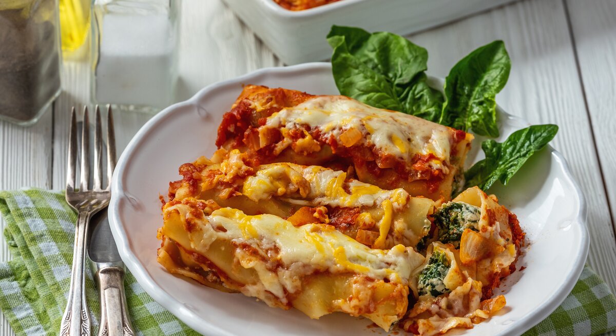 Weißer Teller belegt mit Cannelloni mit Ricotta-Spinat-Füllung und Tomatensoße auf hölzernem Untergrund.