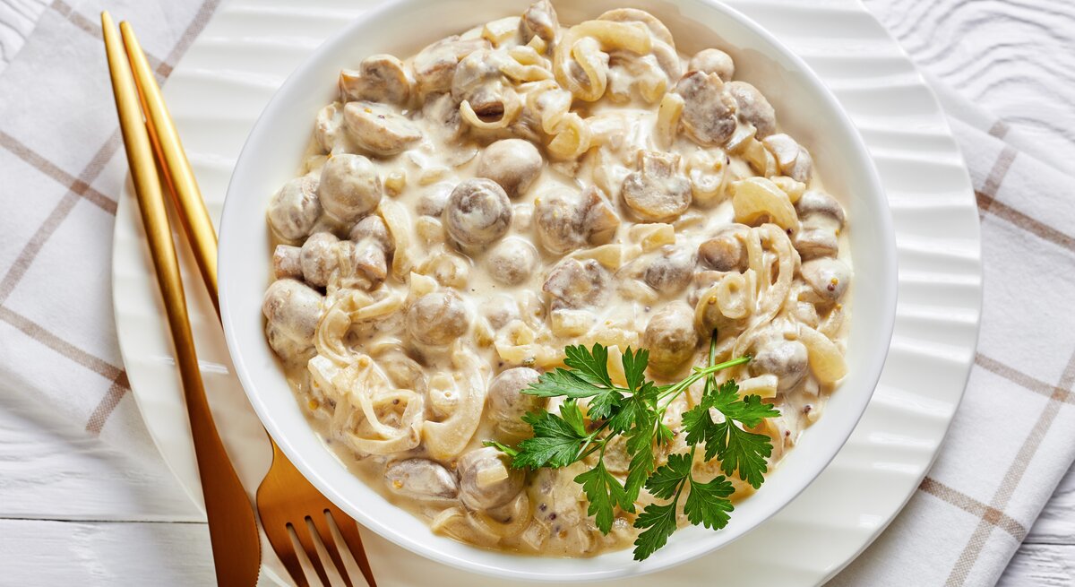Rahmchampignons mit Nudeln auf weißem Teller, angerichtet auf einem hellen Tischtuch und zu Holzbesteck.