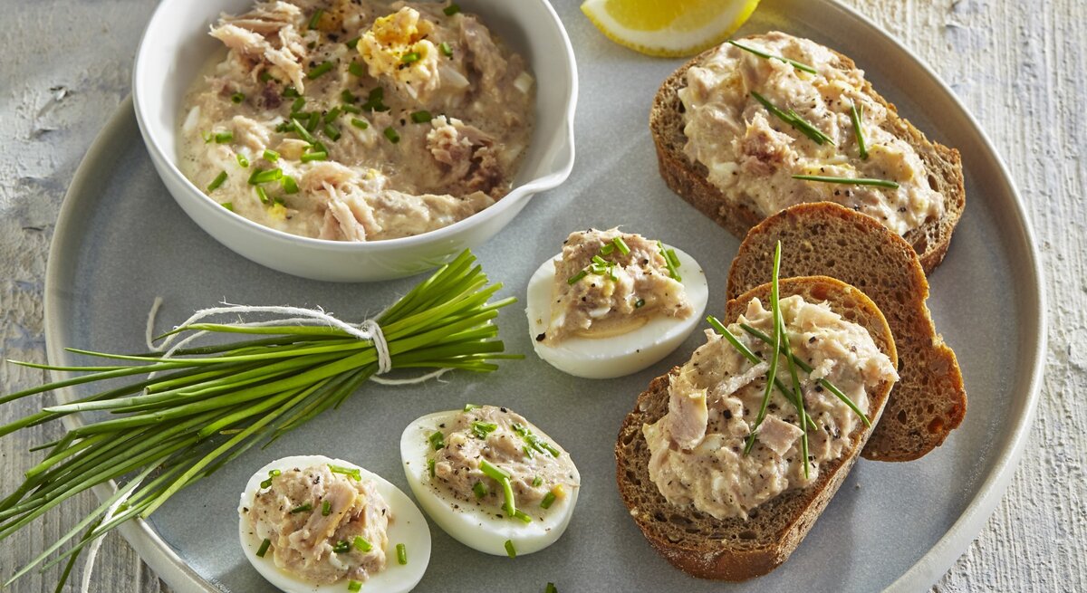 Thunfisch-Ei-Aufstrich in einer Schale und auf Brot und Ei. Daneben liegt Schnittlauch und geschnittene Zitrone. 
