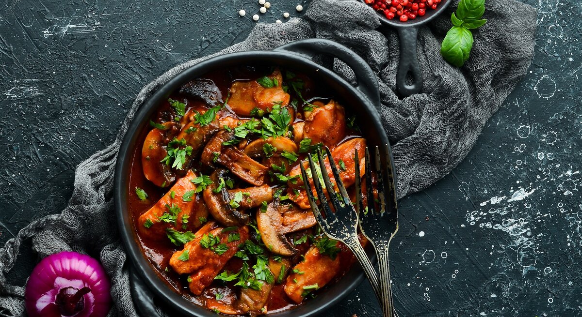 Eine schwarze Schale mit dem Blitz-Gulasch auf einem dunklen Tisch mit rotem Pfeffer und Basilikum im Hintergrund.