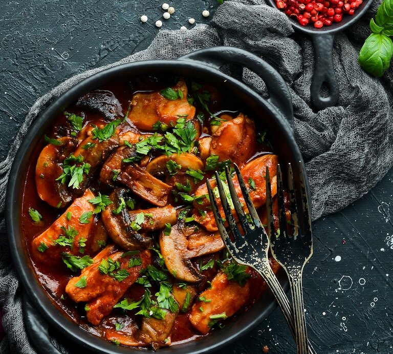 Eine schwarze Schale mit dem Blitz-Gulasch auf einem dunklen Tisch mit rotem Pfeffer und Basilikum im Hintergrund. | © Shutterstock/ YARUNIV Studio