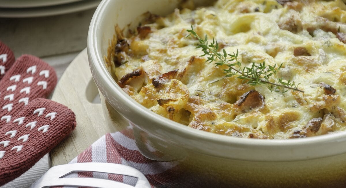 Kretanische Kartoffeln in einer hellen Auflaufform mit frischem Kraut neben Ofenhandschuhen und einem Pfannenwender auf einem hellen Holzbrett neben weiteren Tellern auf einem hellen Tisch. 