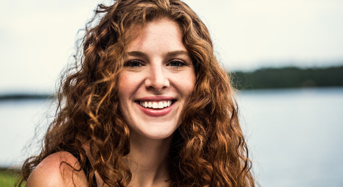 Junge Frau mit langen, lockigen roten Haaren, die in die Kamera lächelt. Sie hat einen frischen, natürlichen Look und strahlt Freude und Zufriedenheit aus.
