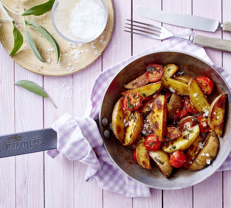 Eine gusseiserne Pfanne mit mediterranen Kartoffeln drinnen, die auf einem rosa karierten Geschirrtuch liegt neben Besteck, Salbei und grobem Meersalz | © EAT CLUB/Isabell Triemer
