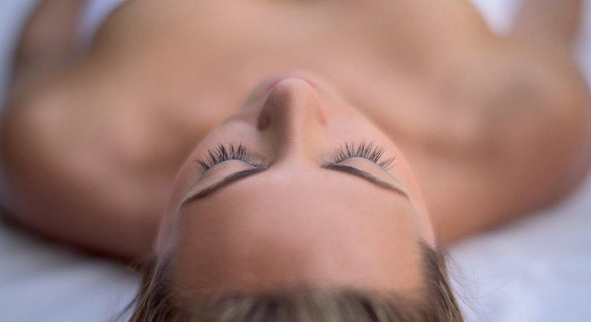 Nahaufnahme des Gesichts einer liegenden Person, die die Augen geschlossen hat. Die Augenlider sind entspannt und die langen, geschwungenen Wimpern sind deutlich zu sehen. Der Fokus liegt auf den Wimpern, die gegen einen unscharfen Hintergrund hervorstechen, welcher die Konturen des Gesichts und des Oberkörpers der Person zeigt."