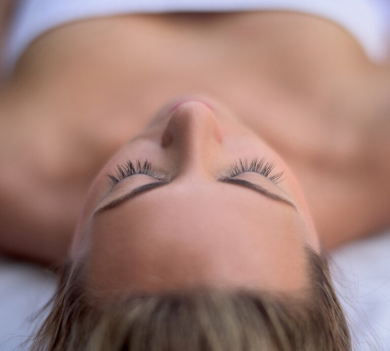 Nahaufnahme des Gesichts einer liegenden Person, die die Augen geschlossen hat. Die Augenlider sind entspannt und die langen, geschwungenen Wimpern sind deutlich zu sehen. Der Fokus liegt auf den Wimpern, die gegen einen unscharfen Hintergrund hervorstechen, welcher die Konturen des Gesichts und des Oberkörpers der Person zeigt." | © iStock/andresr
