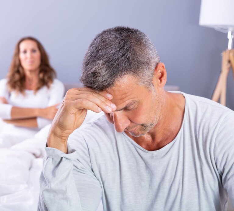 Eine Frau liegt im Bett, ihr Partner sitzt geknickt am Bettrand und schaut nach unten. | © Shutterstock / Andrey_Popov