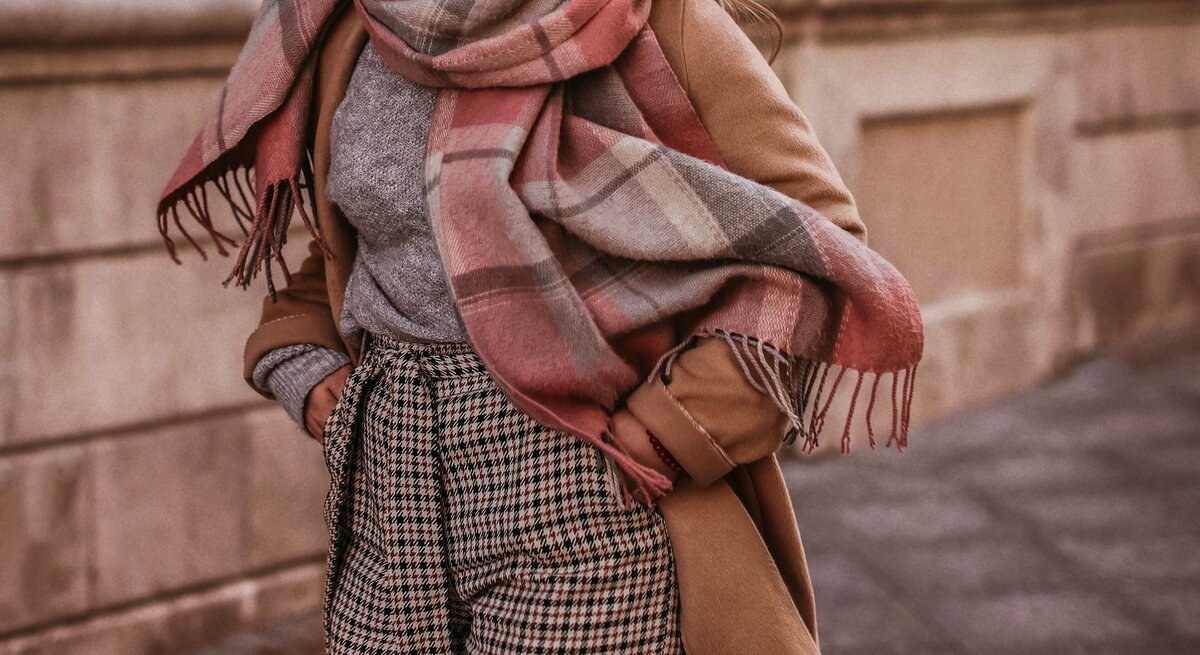 Eine Frau trägt angesagte Strickmode für den Herbst, wie eine karrierte Hose, einen graumelierten Pullover und einen XXL-Schal in braun, rosa und weiß. Dazu kombiniert sie einen hellbraunen Mantel während sie durch die Straßen läuft.