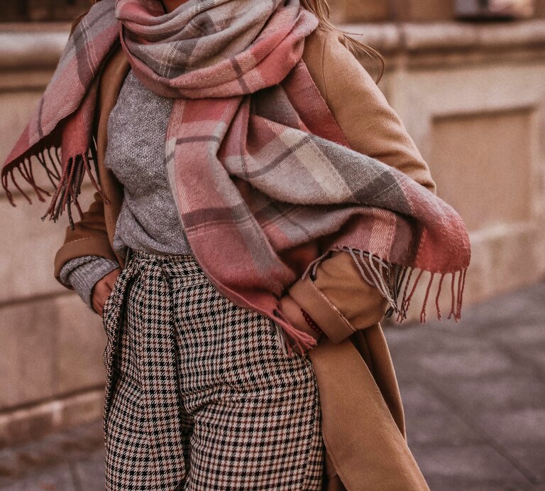Eine Frau trägt angesagte Strickmode für den Herbst, wie eine karrierte Hose, einen graumelierten Pullover und einen XXL-Schal in braun, rosa und weiß. Dazu kombiniert sie einen hellbraunen Mantel während sie durch die Straßen läuft. | © Adobe Stock/ asauriet