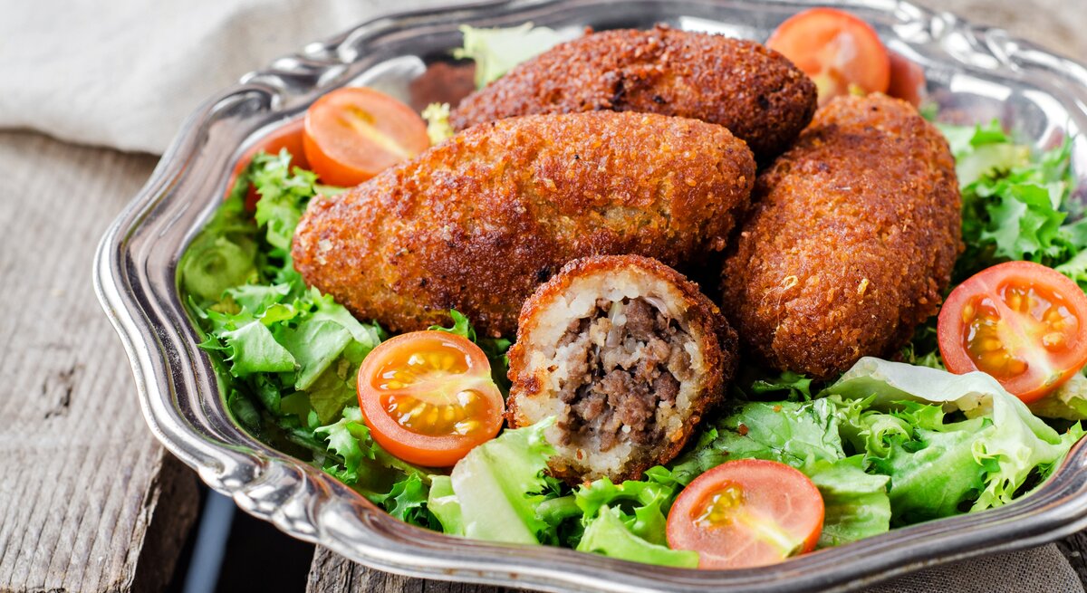 Schräge Frontalsicht: Auf einem runden silbernen Teller liegen Kibbeh, orienatlische Bulgur-Bällchen, die mit Hackfleisch gefüllt sind. Drum sind grüne Salatblätter und halbierte roten Tomaten.