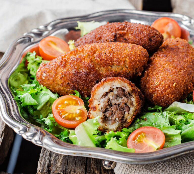 Schräge Frontalsicht: Auf einem runden silbernen Teller liegen Kibbeh, orienatlische Bulgur-Bällchen, die mit Hackfleisch gefüllt sind. Drum sind grüne Salatblätter und halbierte roten Tomaten. | © Shutterstock / Tanya Stolyarevskaya