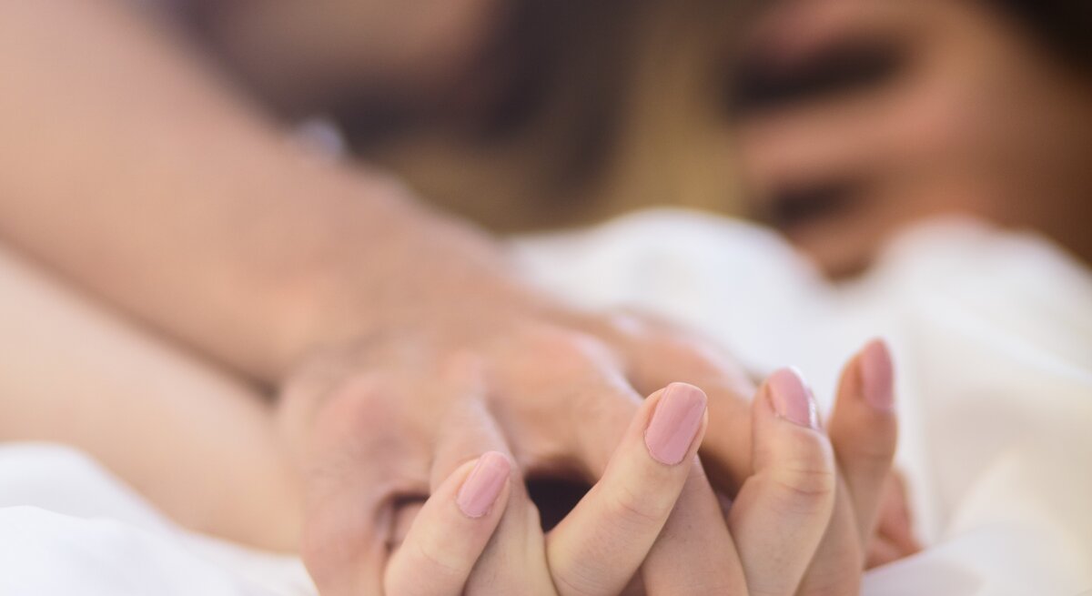 Hände eines Paares, die sich festhalten, im Vordergrund, mit unscharfen Gesichtern im Hintergrund, was Intimität andeutet.