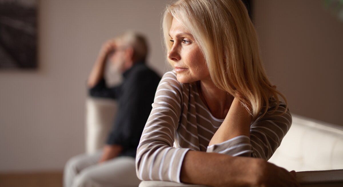 Frau mittleren Alters mit blonden Haaren, die nachdenklich zur Seite schaut. Im Hintergrund ist ein Mann zu sehen, der ebenfalls nachdenklich wirkt und in die entgegengesetzte Richtung schaut. Beide scheinen in einem Wohnzimmer zu sein.