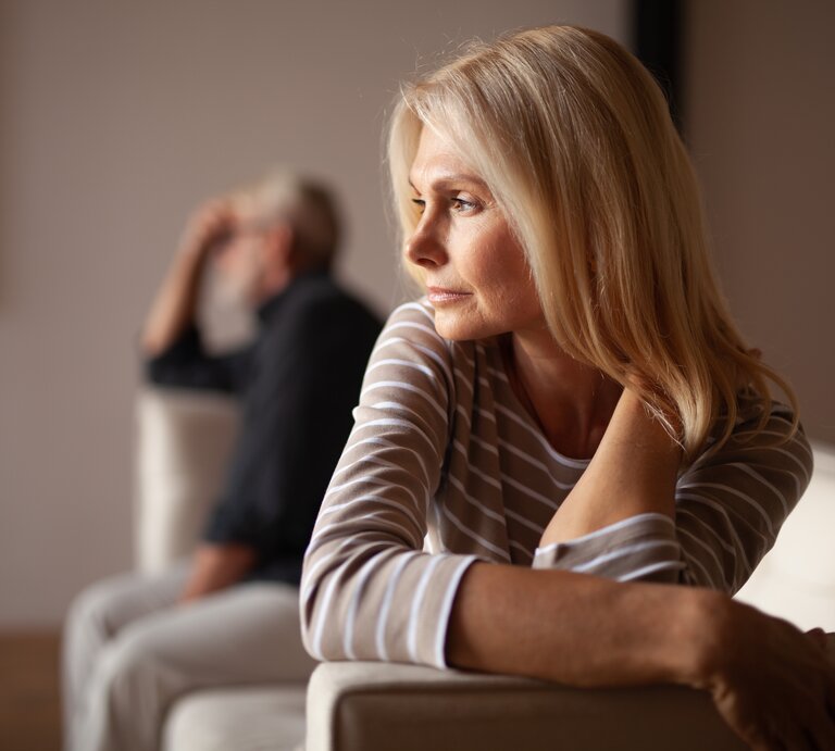 Frau mittleren Alters mit blonden Haaren, die nachdenklich zur Seite schaut. Im Hintergrund ist ein Mann zu sehen, der ebenfalls nachdenklich wirkt und in die entgegengesetzte Richtung schaut. Beide scheinen in einem Wohnzimmer zu sein. | © Getty Images /	Andrii Zastrozhnov