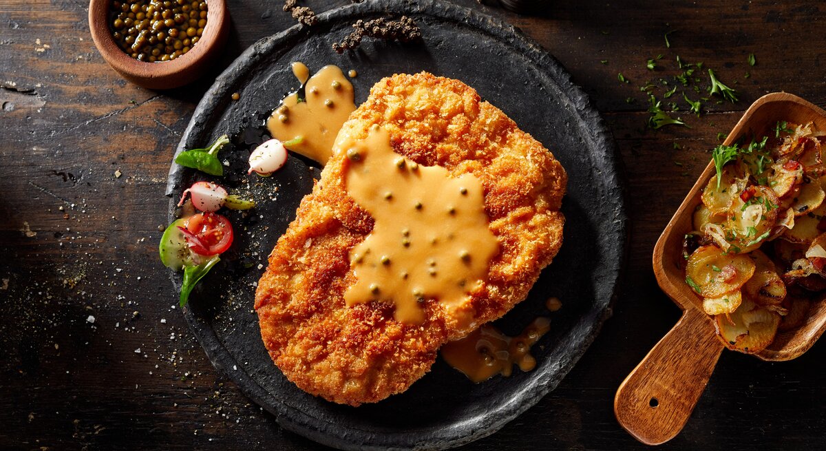 Ein Schwarzer Teller mit dem Schnitzel und der Senfsoße auf einem rustikalem Untergrund.