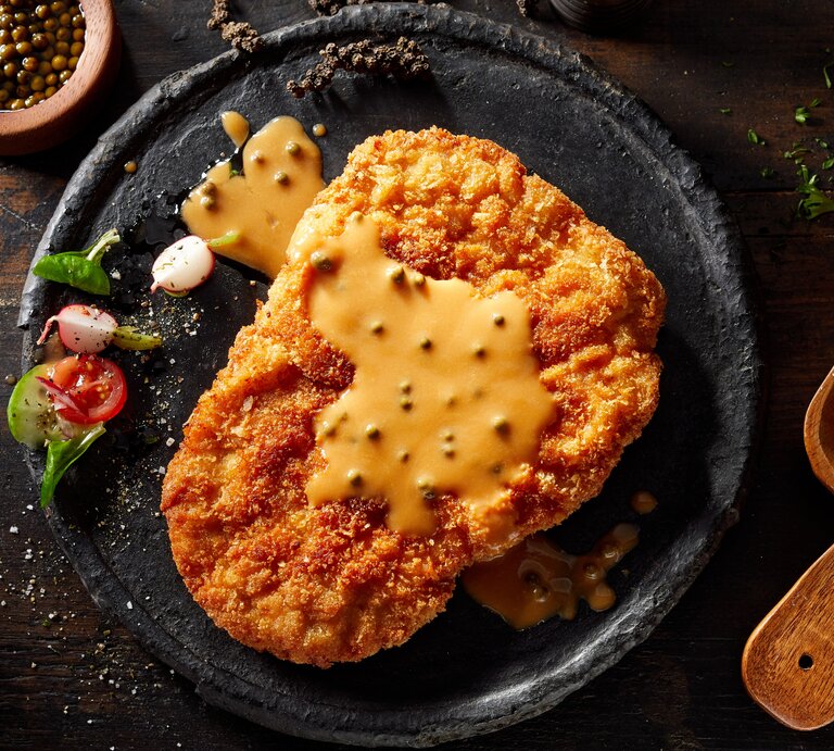 Ein Schwarzer Teller mit dem Schnitzel und der Senfsoße auf einem rustikalem Untergrund. | © Shutterstock/ stockcreations