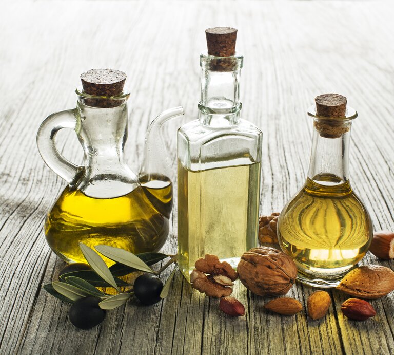 Drei Glasflaschen mit Olivenöl und Essig umgeben von Oliven und Nüssen auf rustikalem Holztisch. | © iStock/dulezidar