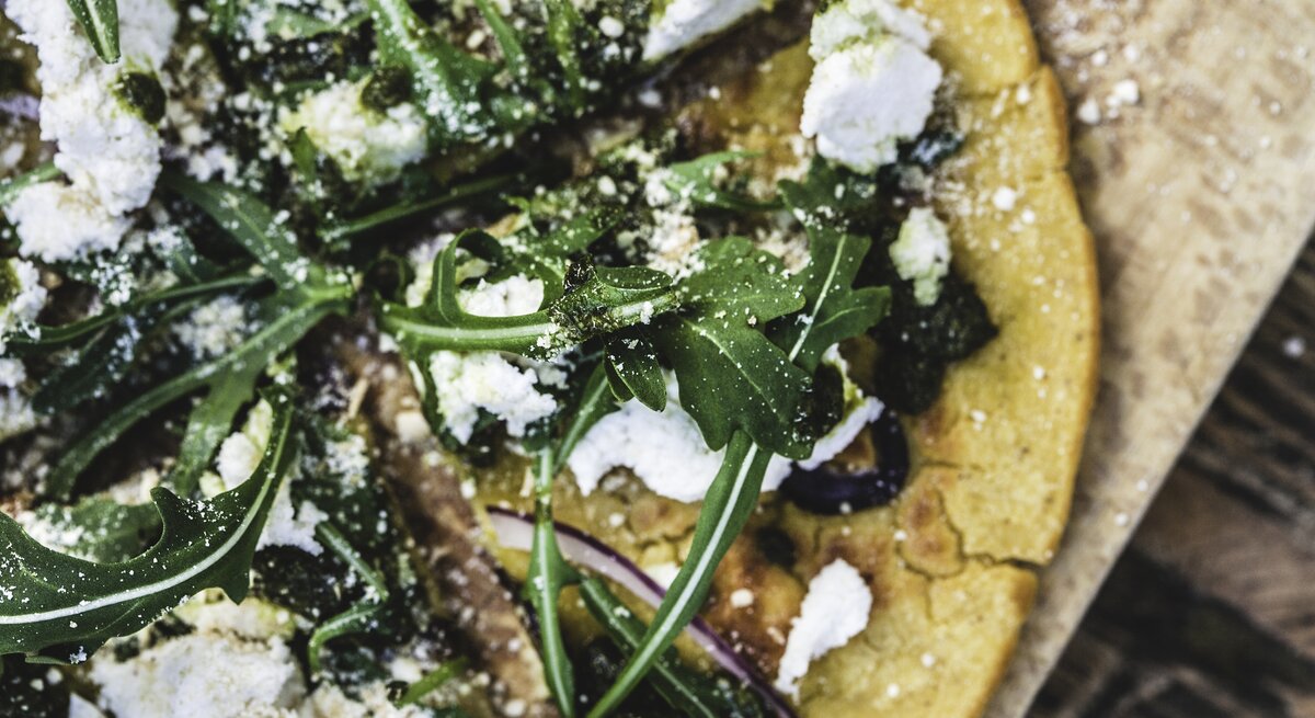 Auf einem Holzbrett ist ein Teil einer Pizza zu sehen. Der Belag besteht aus Feta-Käse, Rucola und roten Zwiebelringen. Der Pizzaboden besteht aus Haferflocken und ist leicht gelblich.