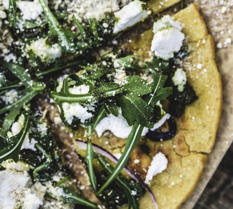 Auf einem Holzbrett ist ein Teil einer Pizza zu sehen. Der Belag besteht aus Feta-Käse, Rucola und roten Zwiebelringen. Der Pizzaboden besteht aus Haferflocken und ist leicht gelblich. | © Enrique Díaz / 7cero
