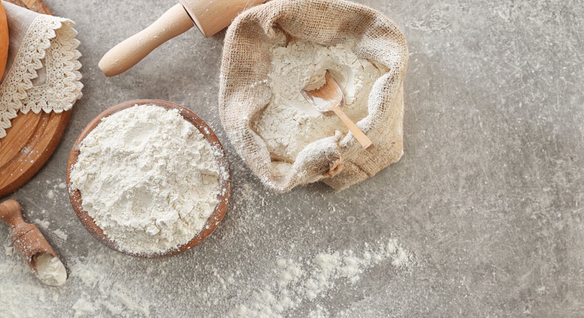 Draufsicht: Auf einem grauen Untergrund stehen ein Leinensack und eine Schüssel mit Self Rising Flour. Daneben liegen eine Backrolle sowie ein Brettchen mit einer Spitzendecke.