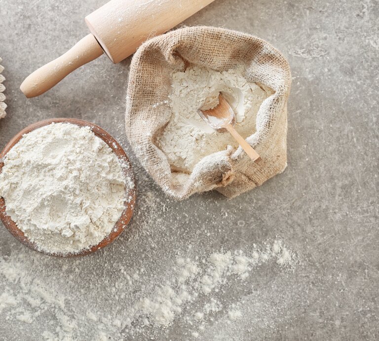 Draufsicht: Auf einem grauen Untergrund stehen ein Leinensack und eine Schüssel mit Self Rising Flour. Daneben liegen eine Backrolle sowie ein Brettchen mit einer Spitzendecke. | © Shutterstock / Africa Studio