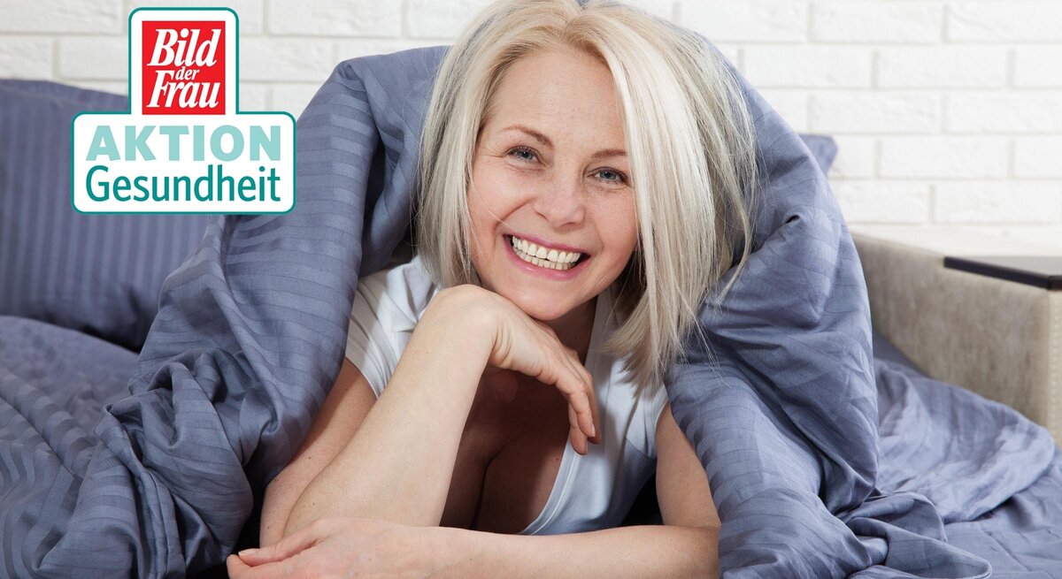 Eine fröhliche Frau mittleren Alters mit strahlendem Lächeln liegt entspannt im Bett, umhüllt von einer grauen Bettdecke. Sie hat kurzes, platinblondes Haar und trägt ein weißes Top, das Frische und Wohlgefühl ausstrahlt. Ihr Kopf ruht auf ihren verschränkten Armen, und ihre Augen sind vor Freude leicht geschlossen. Die weiße Ziegelwand im Hintergrund vermittelt ein modernes, wohnliches Ambiente. In der oberen linken Ecke ist das Logo "Bild der Frau AKTION Gesundheit" zu sehen.