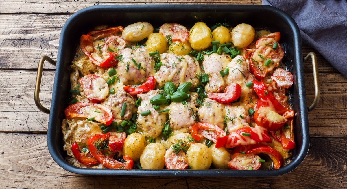 Ein-Blech-Rezept: überbackenes Hähnchen mit Kartoffeln, Paprika, Tomaten und Kräutern in einer Auflaufform, die auf einem Holztisch steht.