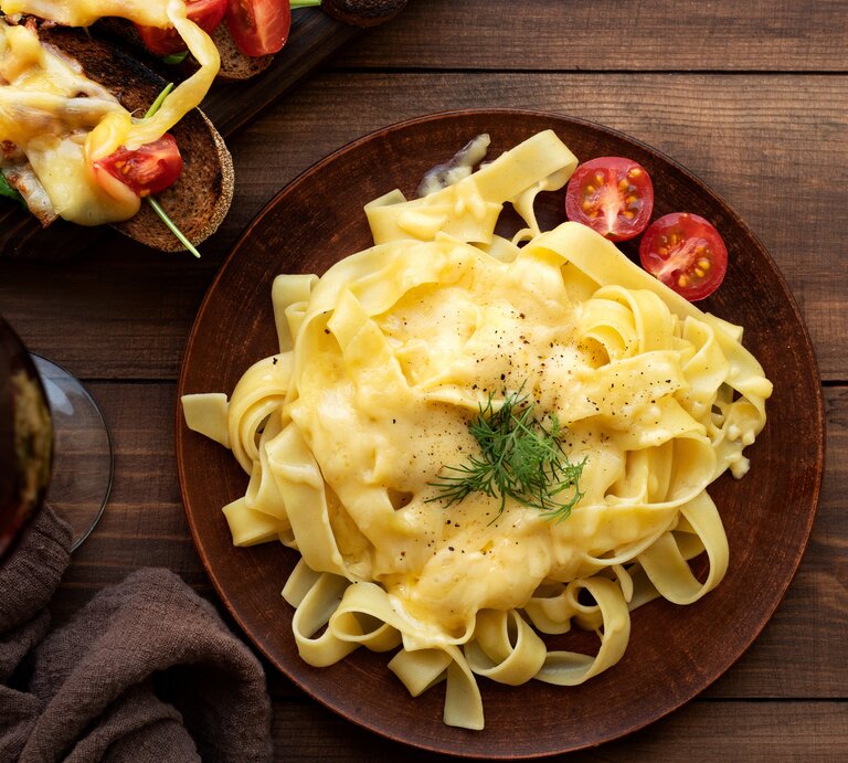 Ein brauner Teller mit Bandnudeln und Käsesoße auf einem Holztisch. Links daneben Raclette-Brote und ein brauner Krug. | © Shutterstock/ Trisha rahman