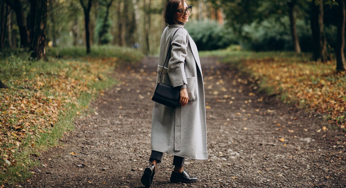 Eine Frau mittleren Alters steht im Herbst im Wald auf einem Waldweg. Sie hat braune Haare und trägt einen grauen Mantel.