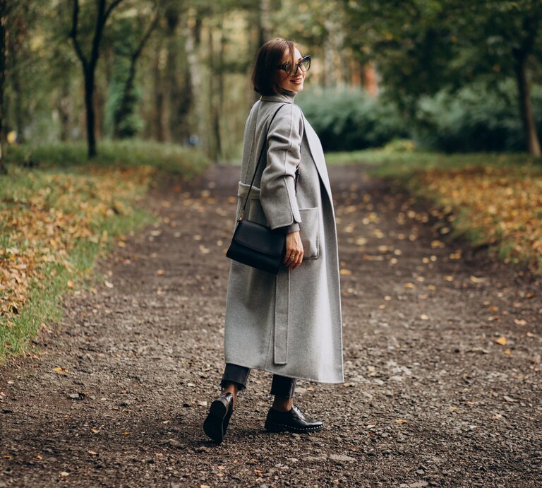 Eine Frau mittleren Alters steht im Herbst im Wald auf einem Waldweg. Sie hat braune Haare und trägt einen grauen Mantel. | © Adobe Stock/ Petro
