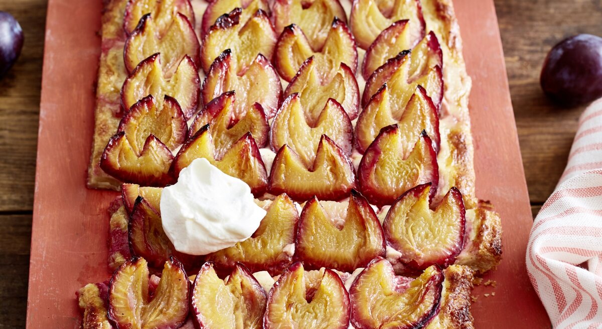 Angeschnittene Pflaumen-Blätterteig-Tarte mit Sahne auf einem blassroten Brett, im Hintergrund Pflaumen und ein Küchentuch, darunter Holz.