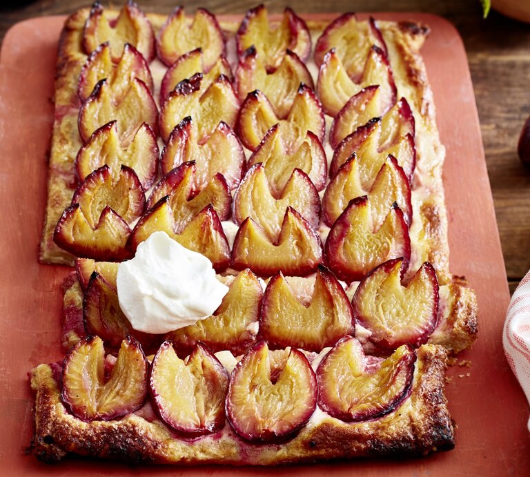 Angeschnittene Pflaumen-Blätterteig-Tarte mit Sahne auf einem blassroten Brett, im Hintergrund Pflaumen und ein Küchentuch, darunter Holz. | © EAT CLUB / Isabell Triemer
