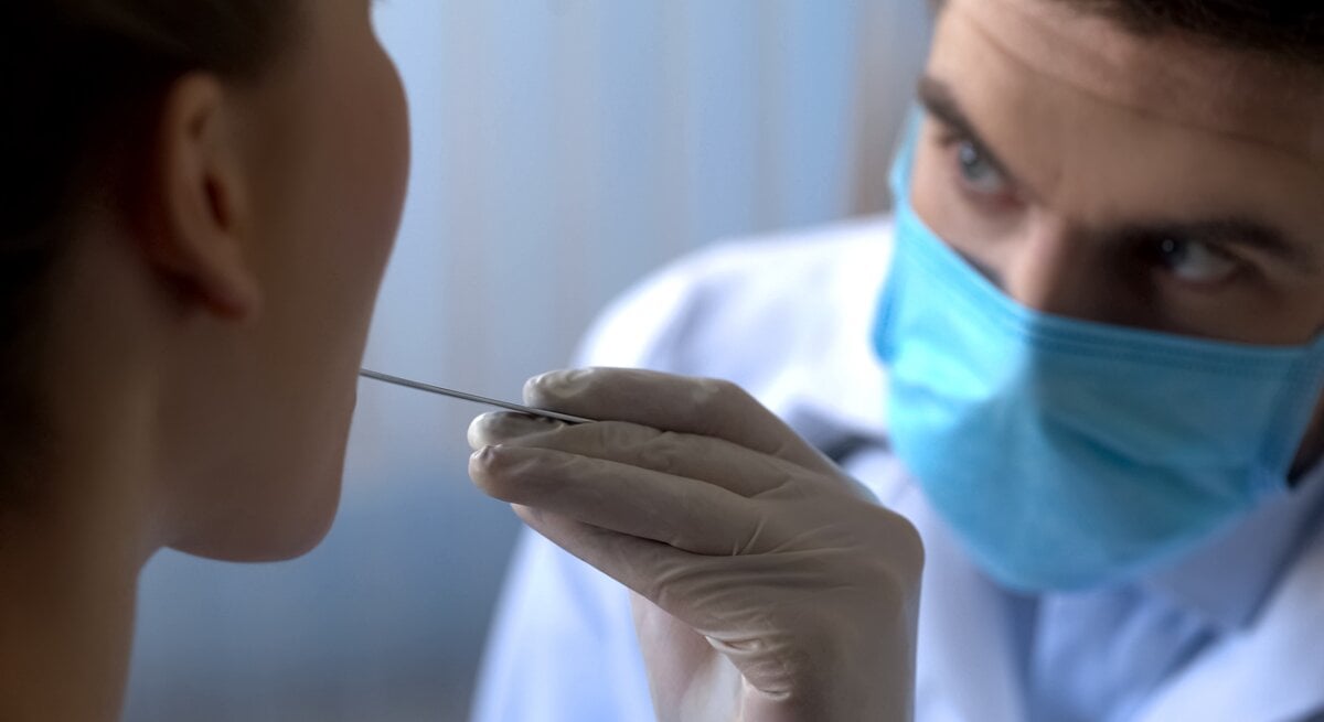 Ein Mediziner in blauer Schutzmaske führt mit Handschuhen einen Mundabstrich bei einer Patientin durch.
