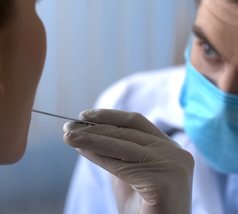 Ein Mediziner in blauer Schutzmaske führt mit Handschuhen einen Mundabstrich bei einer Patientin durch. | © iStock/Motortion