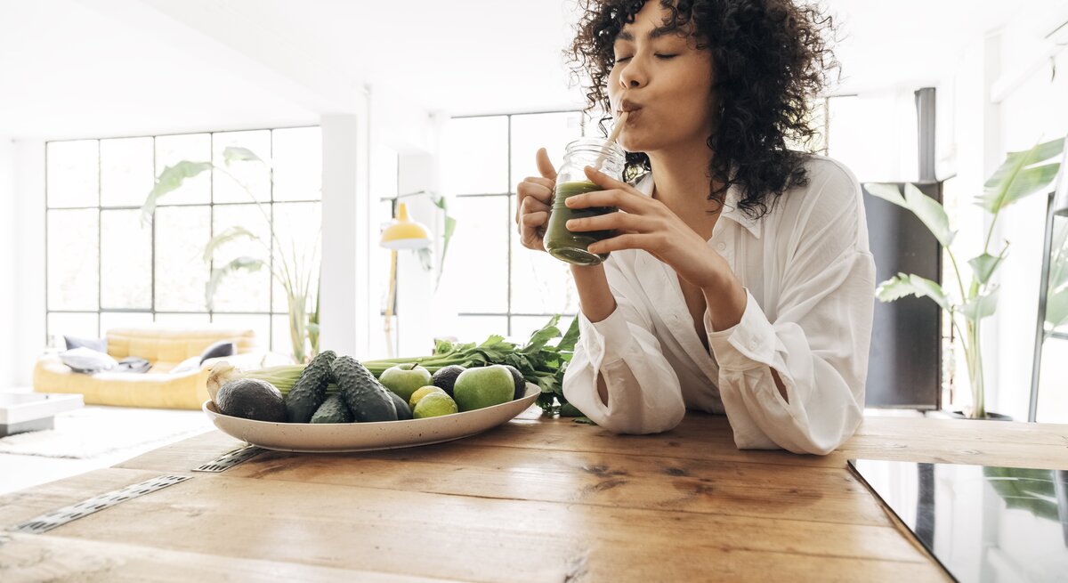 Eine Frau mit lockigem Haar genießt einen grünen Smoothie, während sie an einem Holztisch sitzt. Vor ihr liegt ein Teller mit frischen grünen Früchten und Gemüse wie Avocados und Limetten. Sie trägt eine weiße Bluse und der Raum ist hell und freundlich, mit einem großen Fenster und Pflanzen im Hintergrund, was eine gesunde und entspannte Atmosphäre schafft.