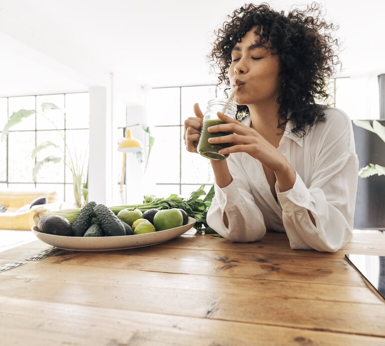 Eine Frau mit lockigem Haar genießt einen grünen Smoothie, während sie an einem Holztisch sitzt. Vor ihr liegt ein Teller mit frischen grünen Früchten und Gemüse wie Avocados und Limetten. Sie trägt eine weiße Bluse und der Raum ist hell und freundlich, mit einem großen Fenster und Pflanzen im Hintergrund, was eine gesunde und entspannte Atmosphäre schafft. | © Getty Images/Daniel de la Hoz