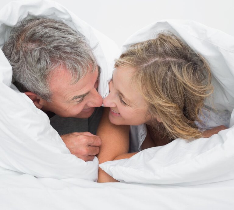 Ein Paar liegt nebeneinander im Bett und schaut sich lächelnd an, die Bettdecke ist über beide Köpfe gezogen | © Shutterstock / wavebreakmedia