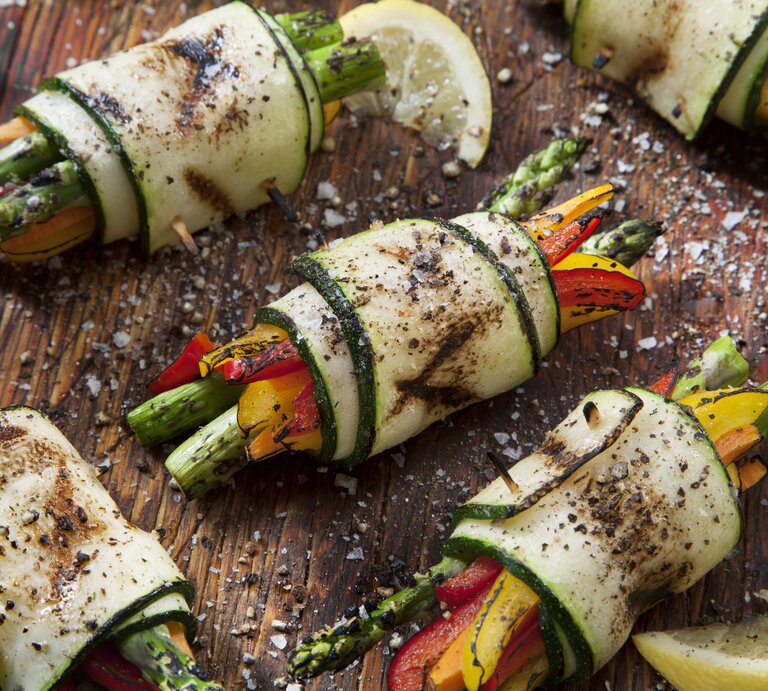Holzbrett belegt mit Zucchini-Röllchen mit Spargel und Paprika, bestreut mit Meersalz und frischem Pfeffer.  | © Getty Images/LauriPatterson