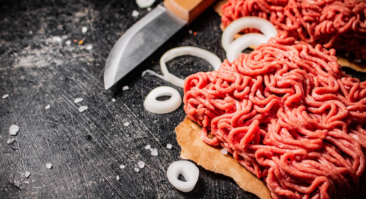 Draufsicht: Rohes Fleisch, das durch den Fleischwolf gedreht wurde liegt auf einem dunklen Untergrund. Daneben liegen rohe Zwiebelringe und ein Küchenmesser.