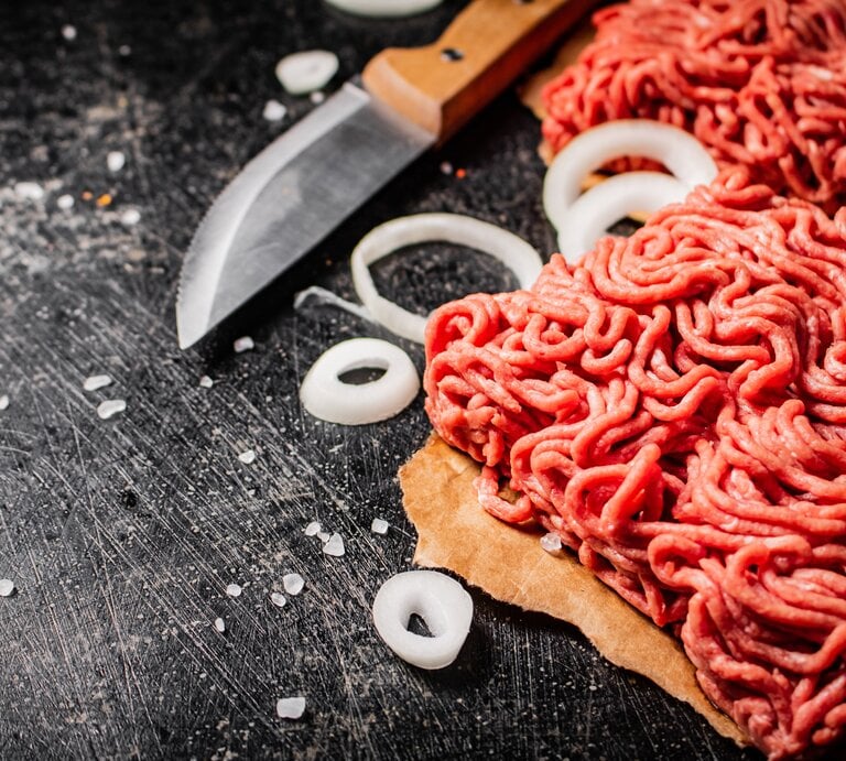 Draufsicht: Rohes Fleisch, das durch den Fleischwolf gedreht wurde liegt auf einem dunklen Untergrund. Daneben liegen rohe Zwiebelringe und ein Küchenmesser. | © Shuttersock / Olesya_sh
