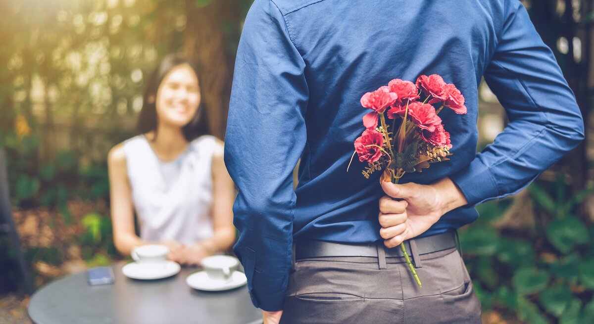 Ein junger Mann hält einen Blumenstrauß hinter seinem Rücken, vor ihm sitzt eine junge Frau und lächelt ihn an