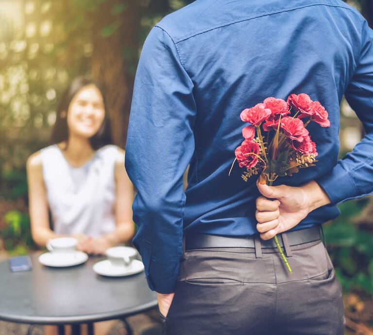 Ein junger Mann hält einen Blumenstrauß hinter seinem Rücken, vor ihm sitzt eine junge Frau und lächelt ihn an | © iStock.com/flukyfluky