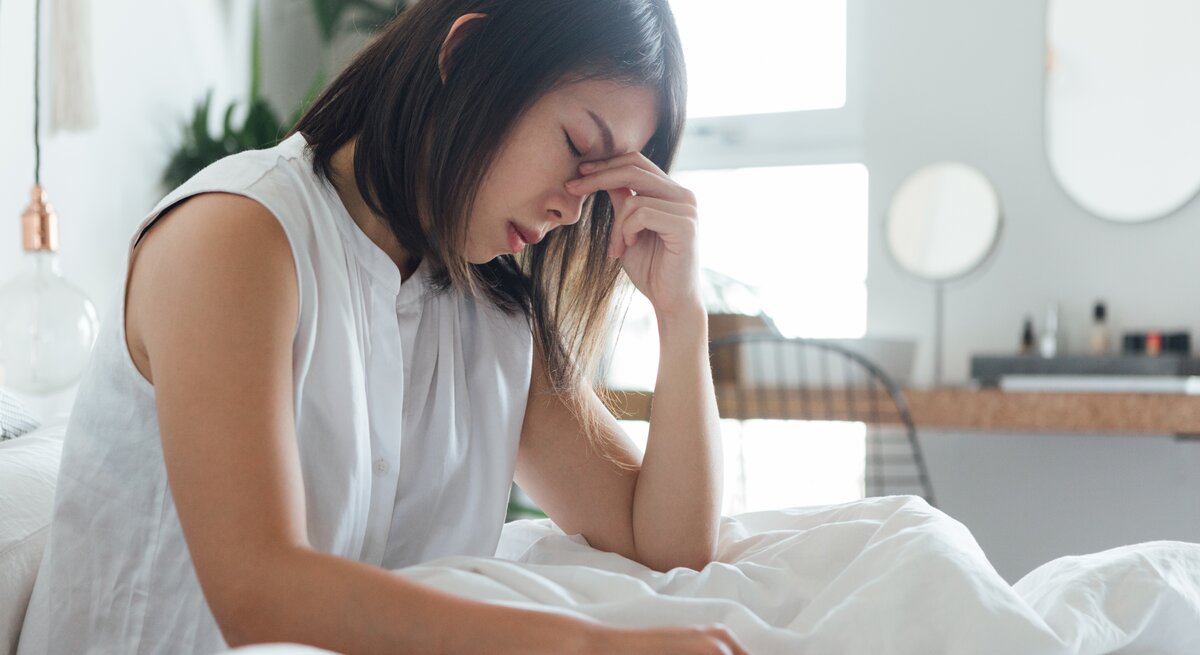  Das Bild zeigt eine junge Frau, die in einem hell erleuchteten, modern eingerichteten Schlafzimmer auf einem Bett sitzt. Sie trägt ein weißes Oberteil und hält mit geschlossenen Augen und einem schmerzverzerrten Gesichtsausdruck ihre Stirn, was darauf hindeutet, dass sie möglicherweise unter Kopfschmerzen oder Stress leidet. Die weiße Bettwäsche und das minimalistische Design des Zimmers sorgen für eine ruhige Atmosphäre, die im Gegensatz zu ihrem offensichtlichen Unbehagen steht.