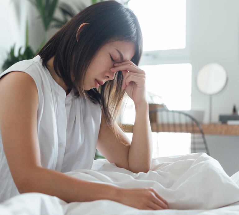  Das Bild zeigt eine junge Frau, die in einem hell erleuchteten, modern eingerichteten Schlafzimmer auf einem Bett sitzt. Sie trägt ein weißes Oberteil und hält mit geschlossenen Augen und einem schmerzverzerrten Gesichtsausdruck ihre Stirn, was darauf hindeutet, dass sie möglicherweise unter Kopfschmerzen oder Stress leidet. Die weiße Bettwäsche und das minimalistische Design des Zimmers sorgen für eine ruhige Atmosphäre, die im Gegensatz zu ihrem offensichtlichen Unbehagen steht. | © Getty Images/Oscar Wong
