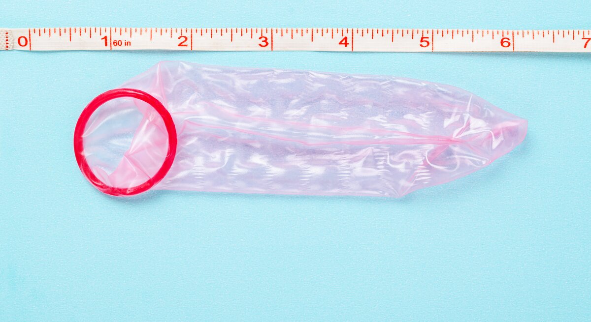 Ein rotes Kondom neben einem Maßband, das Zoll und Zentimeter anzeigt, auf einer hellblauen Oberfläche.
