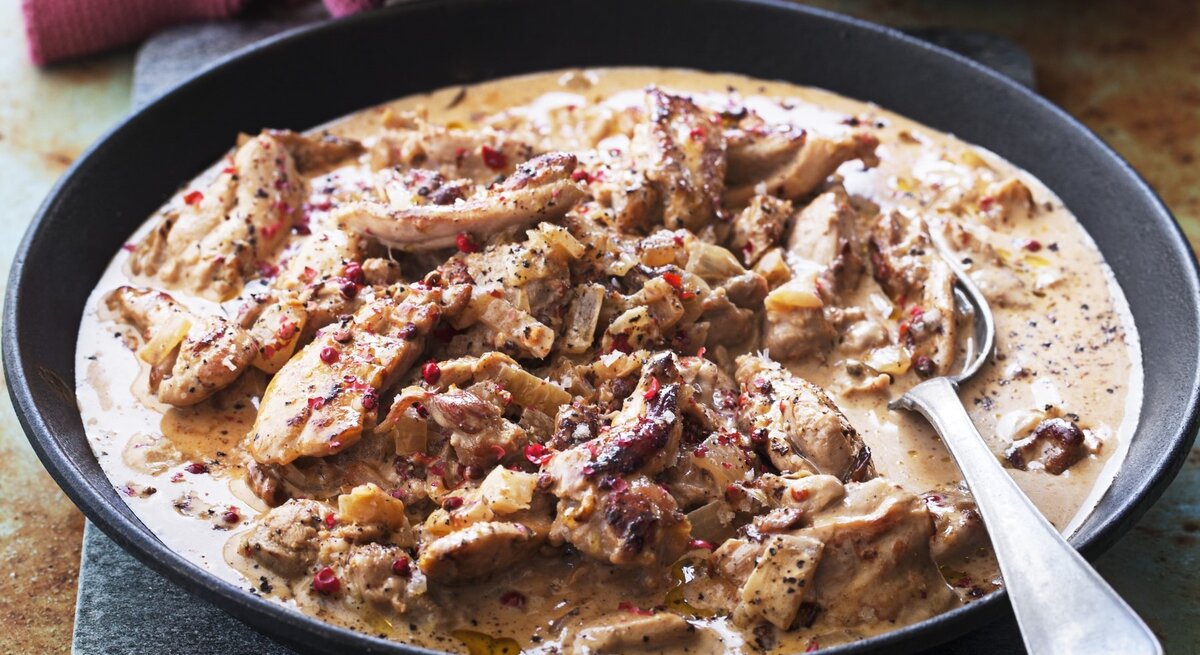 In einer tiefen Pfanne ist eine Portion Hühnereintopf mit Sahne und rosa Pfeffer angerichtet. Sie ist mit roten Pfeffer bestreut.